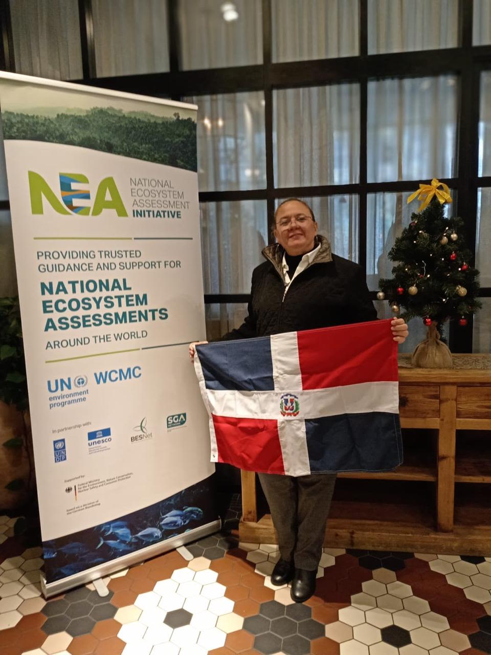 Aleyda Capella holding the Dominican Republic flag next to a National Ecosystem Assessments (NEA) banner at an indoor event