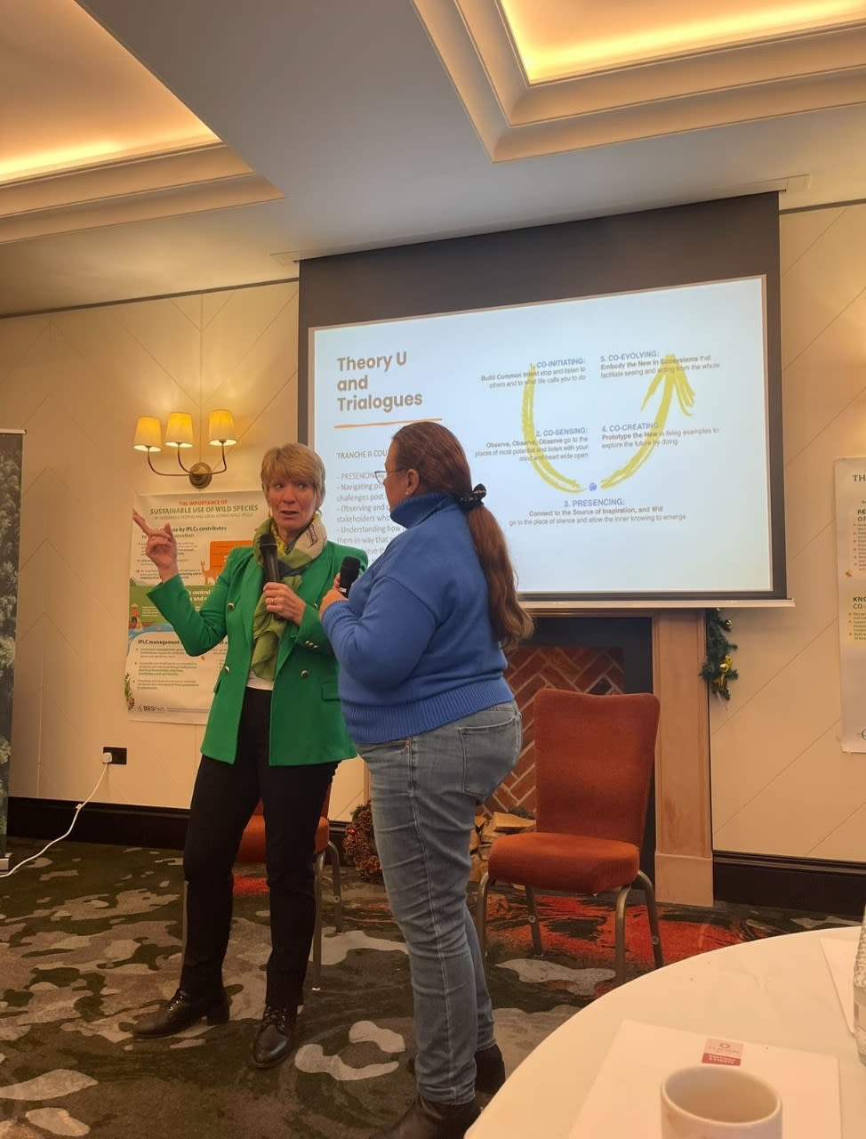 Aleyda Capella, wearing a blue sweater, speaking with another woman in a green blazer during a discussion on Theory U and Trialogues, with a presentation slide visible in the background