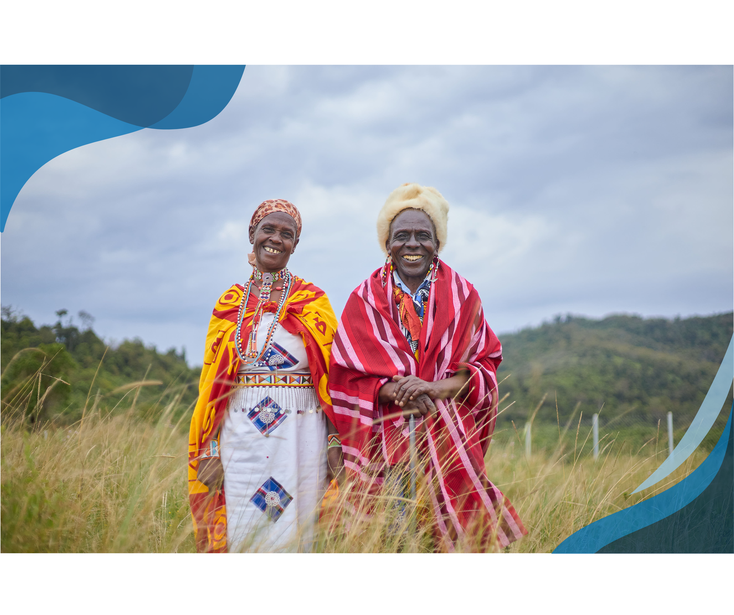 Two smiling individuals dressed in vibrant traditional clothing stand together in a grassy field, with hills and a cloudy sky in the background.