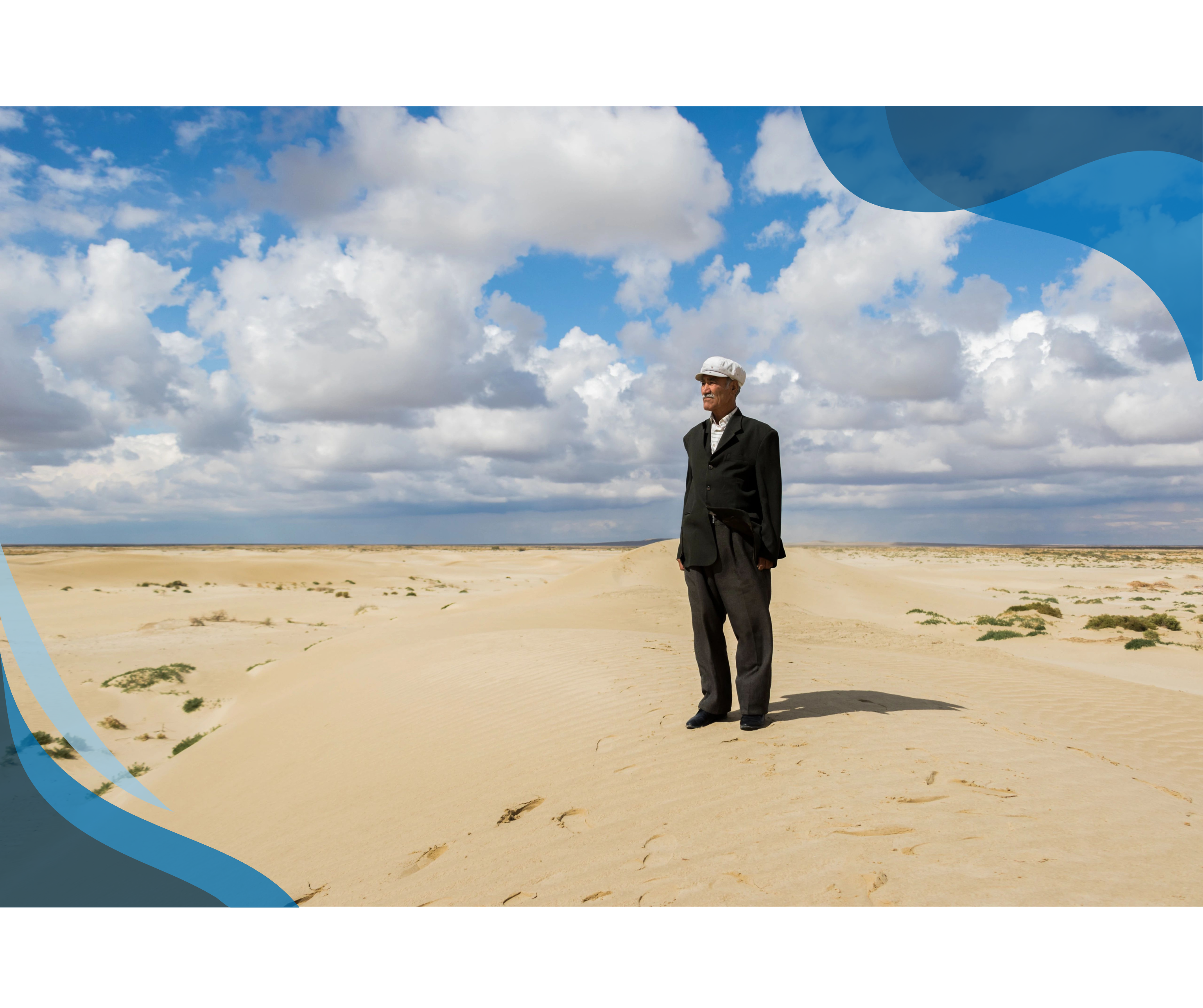  A man in formal attire and a white cap stands alone on a sandy desert landscape under a partly cloudy sky.