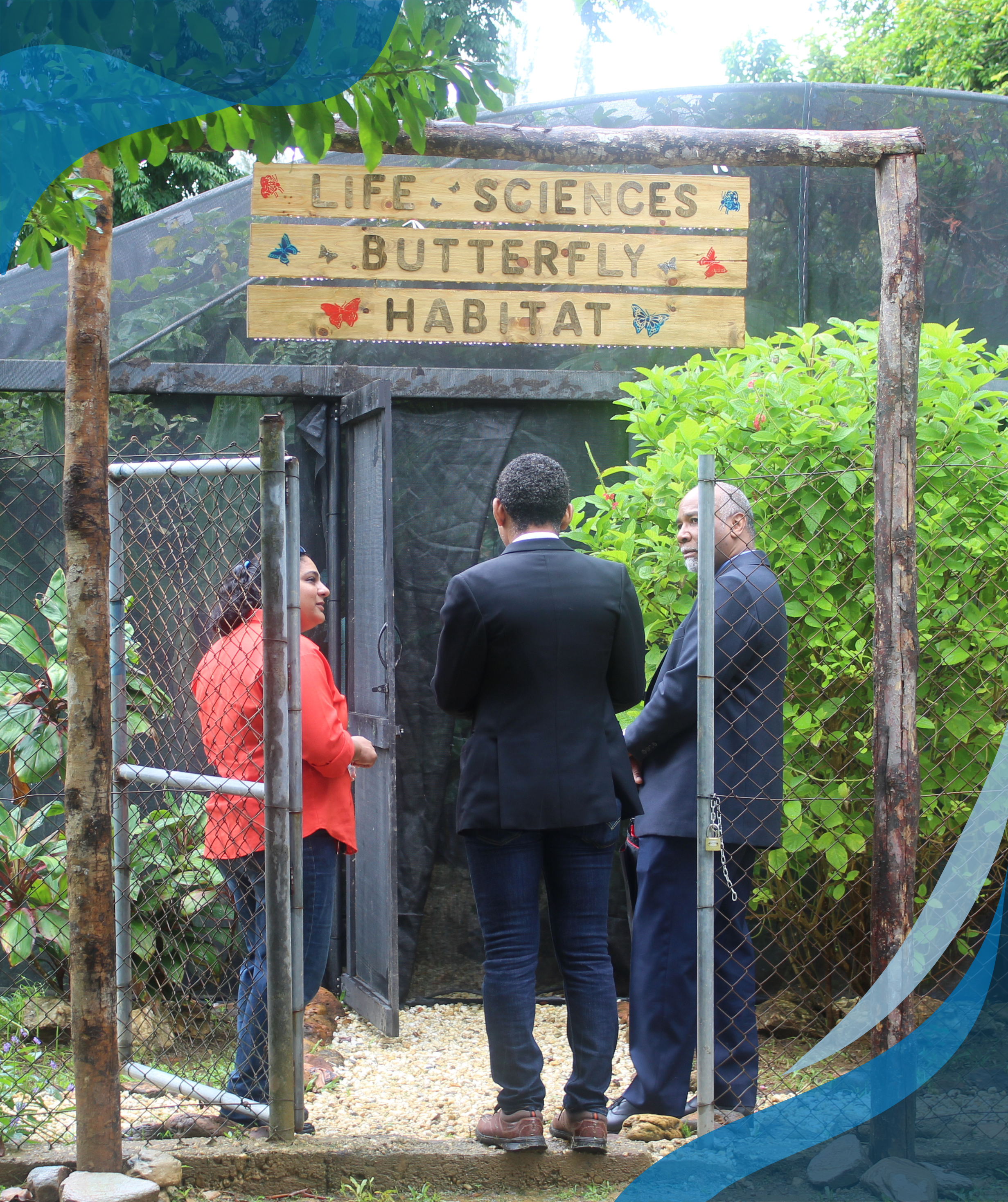 Three people are standing and talking in front of a fenced area labeled "Life Sciences Butterfly Habitat," surrounded by lush greenery.