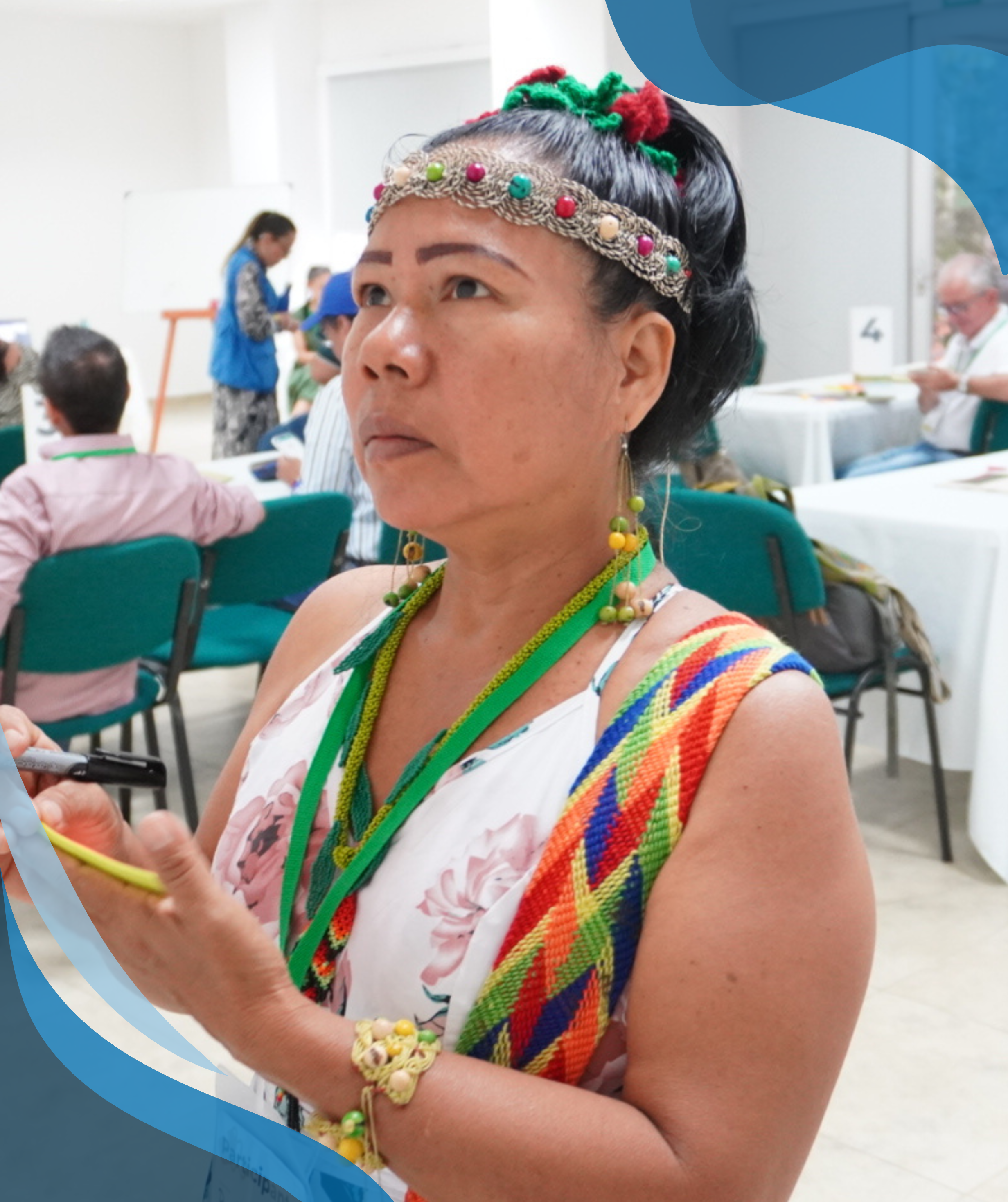 A woman wearing traditional colorful clothing and a headband takes notes while participating in a workshop, with other participants seated at tables in the background.