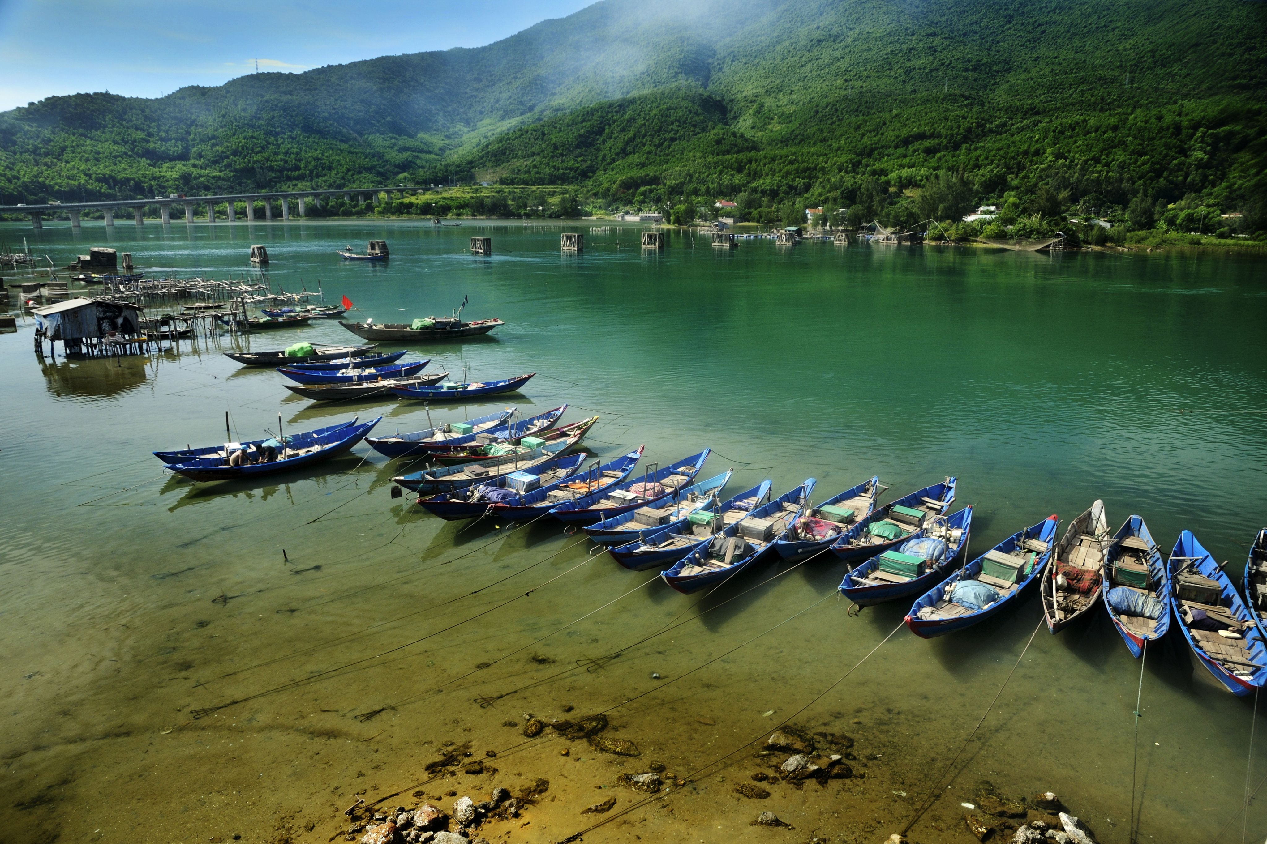 A row of small fishing boats is anchored in the clear, shallow waters of a tranquil lagoon, surrounded by lush green hills and a distant bridge.