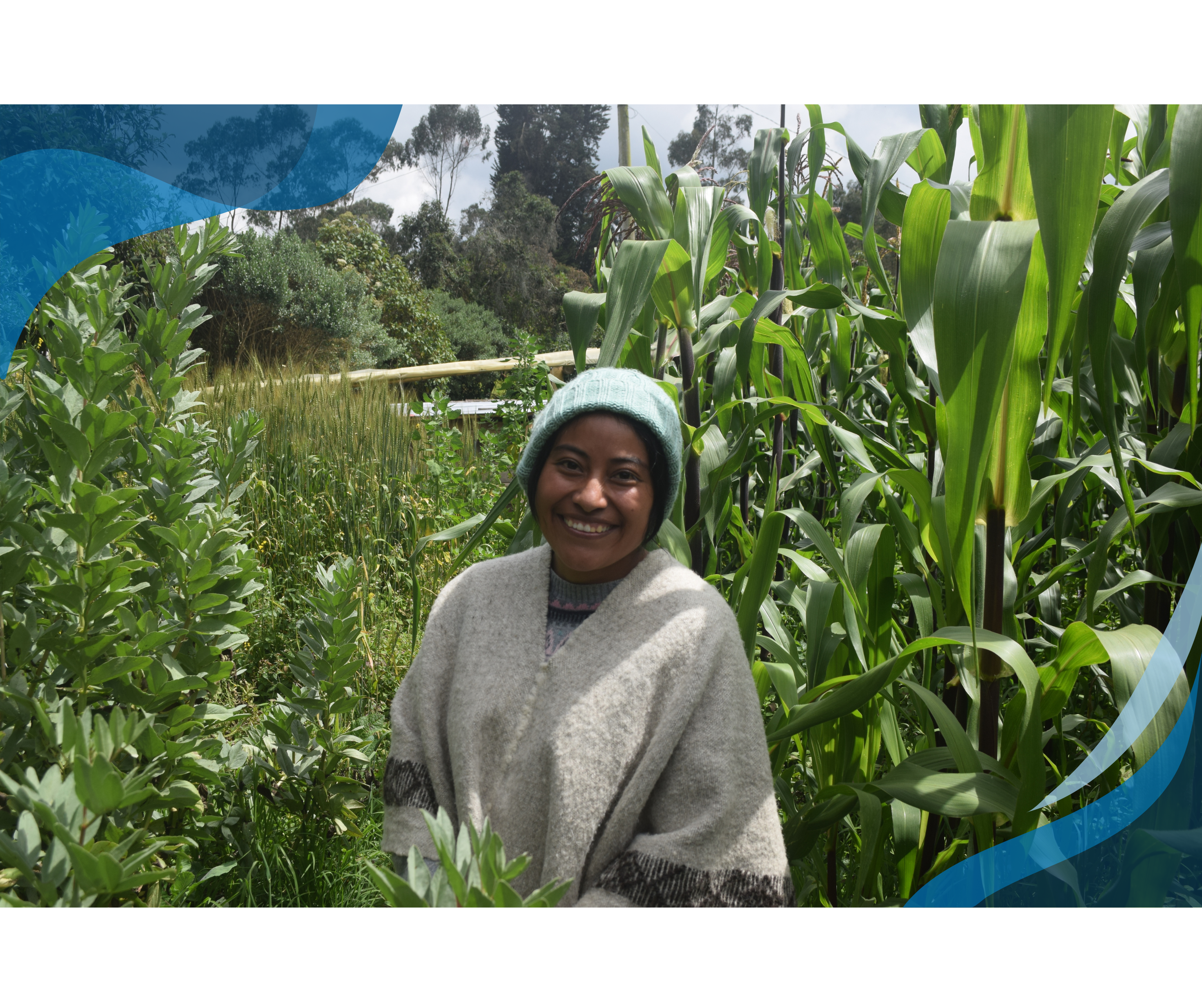 A smiling woman in a woolen poncho and a green knit hat stands amidst tall green plants in a lush field, with trees and mountains in the background.