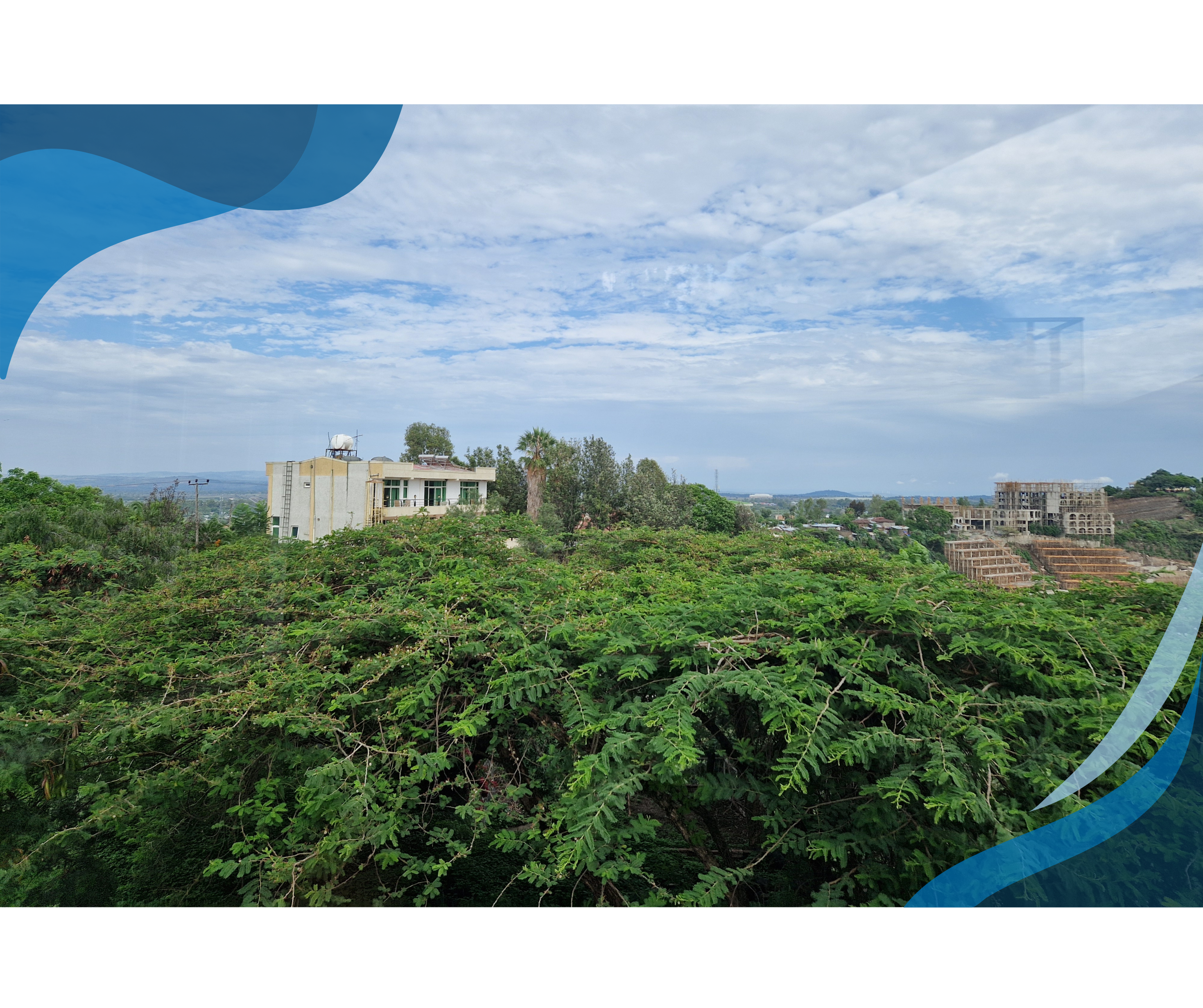 A scenic view of a residential building surrounded by dense green foliage, with partially constructed buildings in the background under a partly cloudy sky.