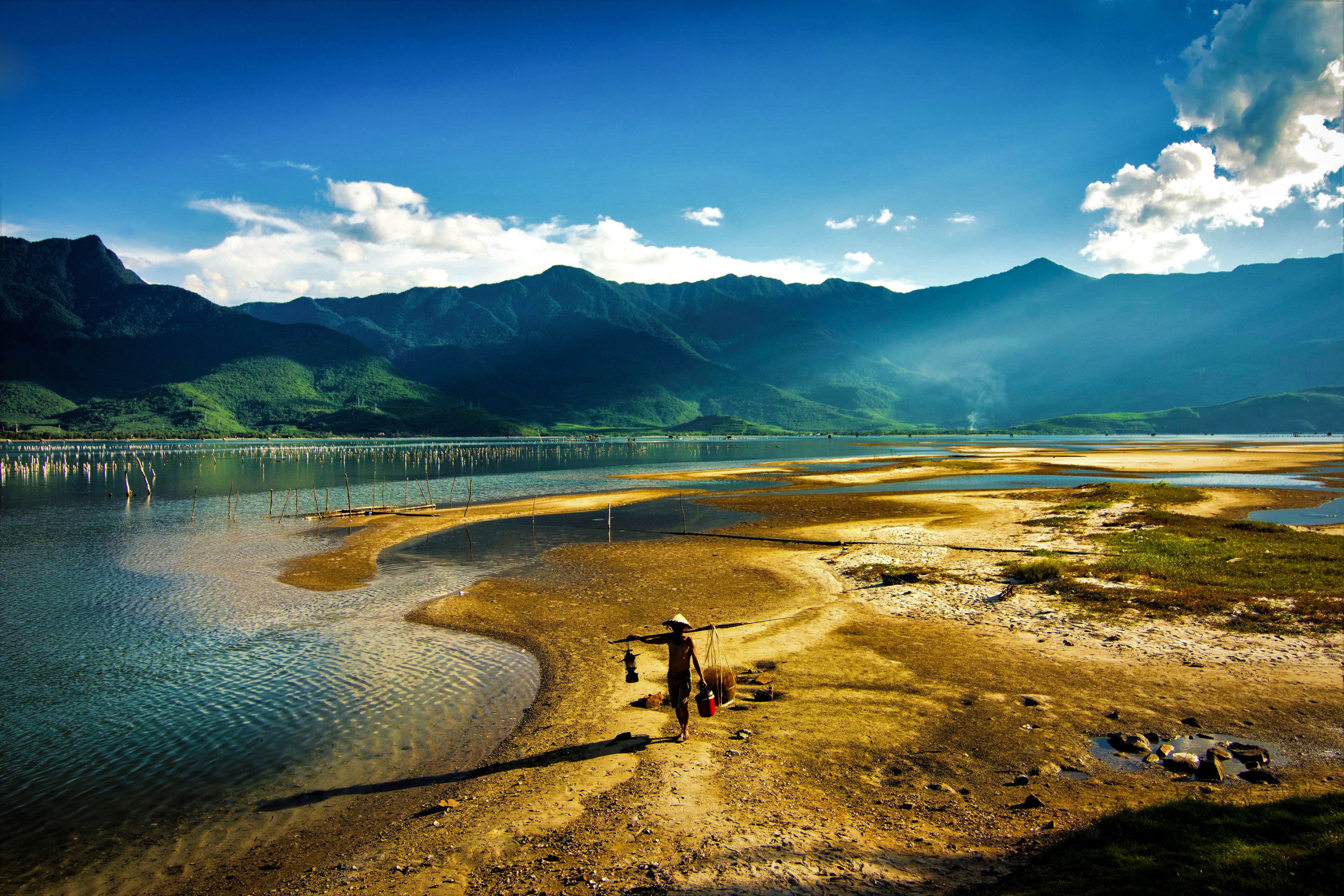 A person wearing a traditional conical hat walks along the sandy shore of a tranquil lagoon, with lush green mountains and a clear blue sky in the background.