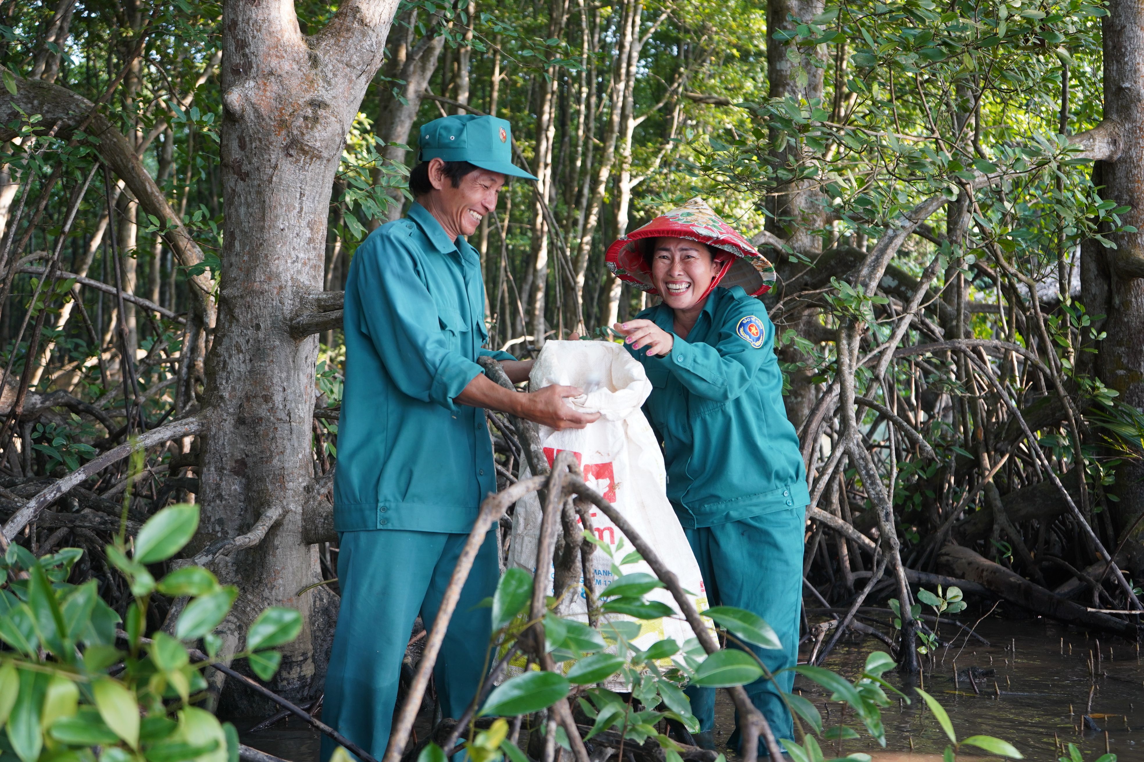  Two people in teal uniforms are smiling and working together in a mangrove forest, holding a large white sack.