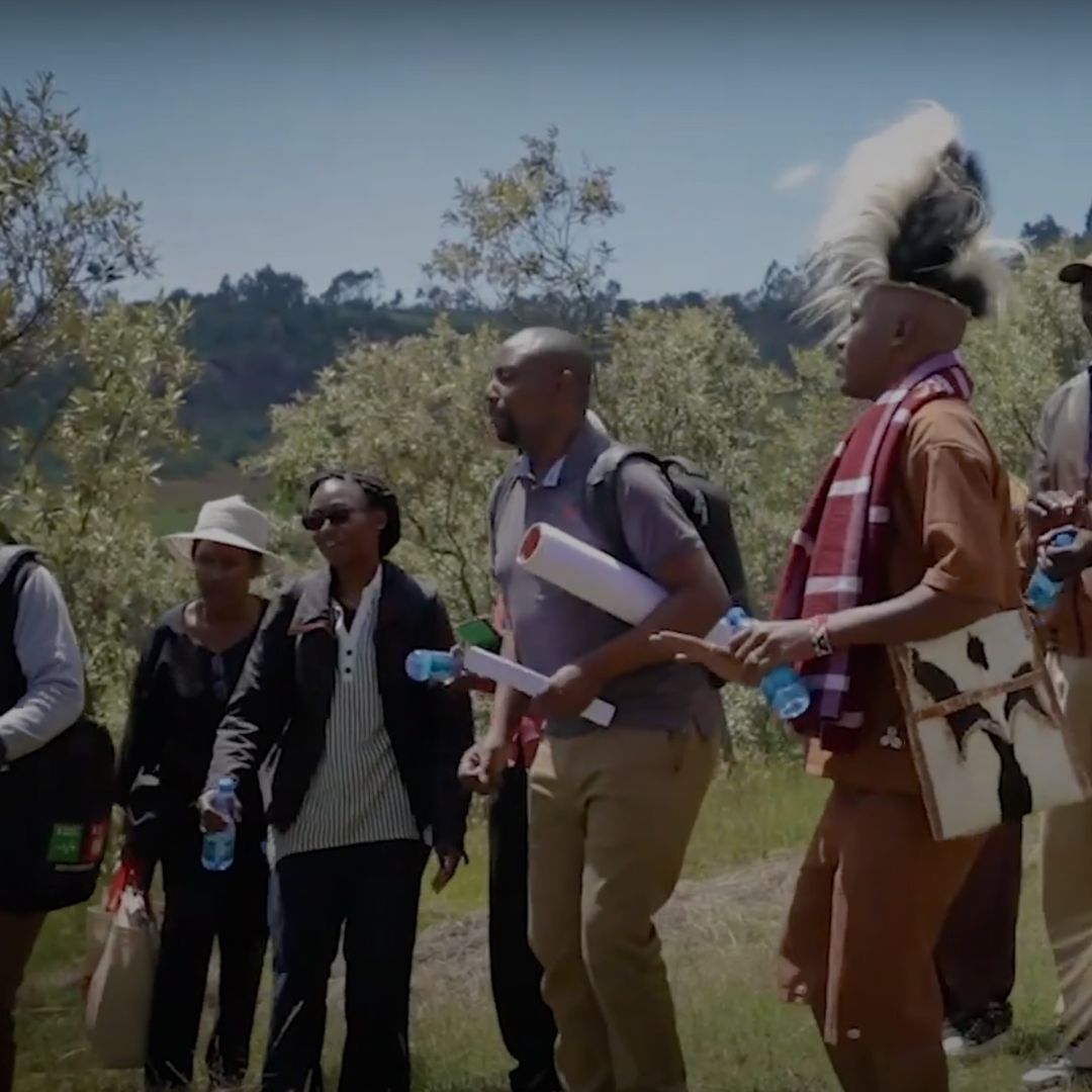 A group of people, including a man in traditional attire, walking outdoors in Eburu, Kenya, carrying water bottles and rolled-up documents.