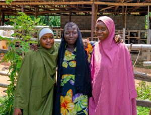 Three young women from a rural community standing close together outdoors and smiling.