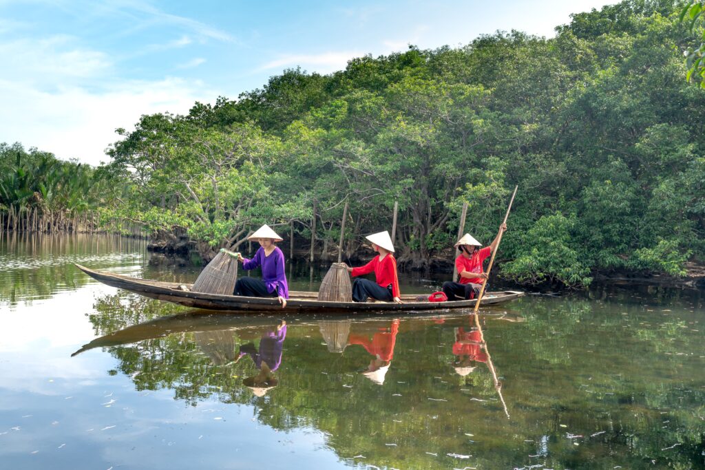Asian fishermen navigate a river in a wooden boat, surrounded by lush mangroves.